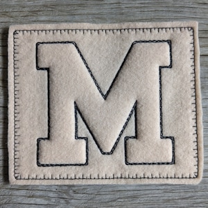Peut inclure: Un écusson en feutre beige avec une bordure cousue noire et une grande lettre "M" noire au centre. L'écusson rectangulaire est posé sur une surface en bois. La couture est visible sur les bords et la lettre.