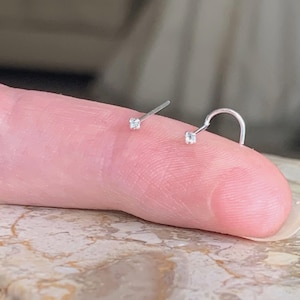 Op de afbeelding: Twee zilveren neusringen met kleine, heldere zirkonia-steentjes. Een ring is een rechte stud en de andere is een gebogen hoepel.