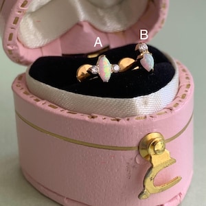 May include: Two gold rings with opal and diamond accents displayed in a pink heart-shaped jewelry box. Ring A features an oval opal with diamond side stones. Ring B has a pear-shaped opal and diamond accents. The box has a gold clasp.