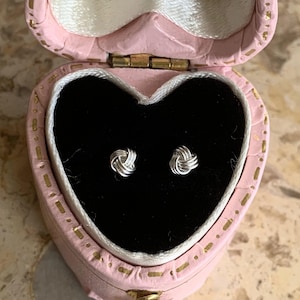 May include: A pair of silver knot-shaped earrings in a pink heart-shaped box with gold trim.