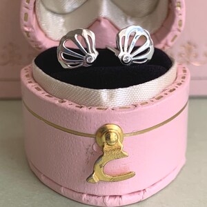 May include: A pair of silver stud earrings shaped like open seashells, each featuring a small, clear, round-cut stone at the base. The earrings are displayed inside a small, oval-shaped, light pink jewellery box with a gold-toned metal latch.