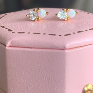 Könnte beinhalten: Ein Paar goldfarbene Ohrstecker. Jeder Ohrring hat einen klaren, runden Edelstein und einen kleineren, ovalen Opal. Die Ohrringe werden auf einer rosa Schmuckschatulle präsentiert.