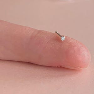 May include: A tiny silver nose stud with a small, clear gemstone. The stud is held on a finger against a soft pink background. The gemstone is round and reflects light.