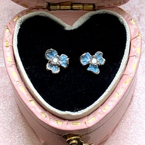 Puede incluir: Un par de pendientes de botón con forma de flor con pétalos de esmalte azul claro y un centro blanco. Los pendientes se muestran dentro de un joyero rosa en forma de corazón con un interior de terciopelo negro.