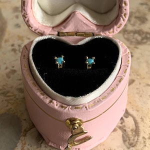 May include: Gold-coloured stud earrings, each with a small, round, turquoise stone, presented in a pink, heart-shaped jewellery box. The box has a gold clasp and trim, and is open to show the earrings on a black velvet interior.