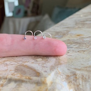 Puede incluir: Tres anillos de nariz de plata con pequeñas piedras preciosas transparentes. Los anillos son curvos y tienen una pequeña bola en el extremo.