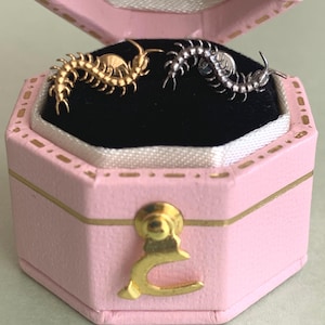 Op de afbeelding: Een paar oorstekers in de vorm van een duizendpoot, één goudkleurig en één zilverkleurig, gepresenteerd in een roze sieradendoosje met gouden accenten. De oorbellen zijn gedetailleerd met gesegmenteerde lichamen en kleine pootjes.