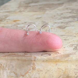 May include: Two silver nose rings with a small clear gemstone on each. The rings are shown on a finger.