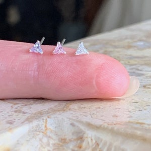 May include: Three small, triangle-shaped stud earrings. The earrings are set with light pink, pink, and clear gemstones. The earrings are displayed on a finger, showcasing their size and design. The earrings have a silver post.