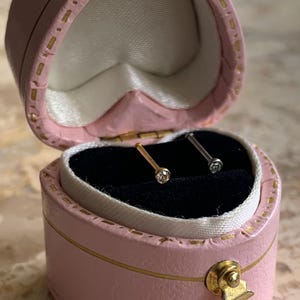 May include: A pair of nose studs displayed in an open, heart-shaped pink jewelry box. One stud is gold-colored, the other silver-colored, each with a small, clear gemstone. The box has gold trim and a gold clasp.
