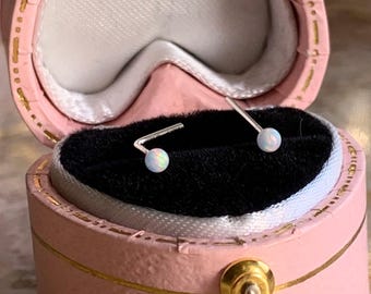 Puede incluir: Un par de pendientes de plata con pequeñas piedras redondas de ópalo blanco. Los pendientes se muestran en una caja de corazón rosa con un cierre dorado.