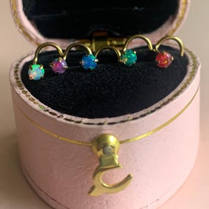 Peut inclure: Collection de cinq piercings de nez avec des pierres d'opale colorées. Chaque anneau est doté d'une barre incurvée dorée. Les anneaux sont présentés dans un écrin rose avec un fermoir doré.