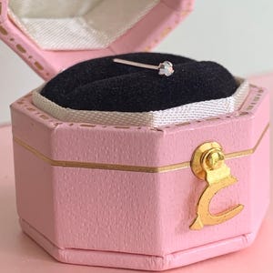 May include: A small earring with a clear gemstone is displayed inside a pink, hexagonal jewellery box. The box has gold accents and a black velvet interior. The earring is set on a thin, silver post.
