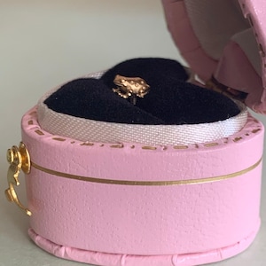 May include: A small gold-toned frog-shaped stud earring with a textured surface, featuring a post back. The earring is displayed inside a round, light pink jewelry box with a gold-colored metal latch and a black velvet interior lining.