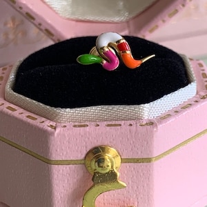 Pode incluir: Um pequeno brinco de pino em metal dourado com formato de verme segmentado. O verme apresenta seções de esmalte em cores vibrantes: branco, vermelho, laranja, rosa e verde. O brinco está apoiado sobre uma almofada de veludo preta dentro de uma caixa de joias rosa.
