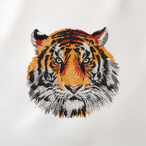May include: Embroidered tiger head with orange, black, and white details. The tiger's face is centered, showcasing its orange and black striped fur, yellow eyes, and pink nose. The embroidery is on a white background.