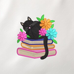May include: An embroidered design featuring a sleeping black cat atop a stack of colorful books. The cat is surrounded by vibrant flowers in shades of orange, pink, and blue, with green leaves. The books have pink, orange, and blue covers.