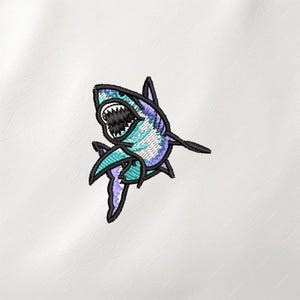 May include: Embroidered shark patch in shades of blue, teal, and purple with black outlines. The shark has an open mouth with visible teeth. The patch is likely for clothing or accessories.