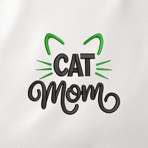 May include: Embroidered design with the words "CAT Mom" in black script. Green cat ears and whiskers are above the word "CAT". The design is on a white background, suitable for clothing or accessories.