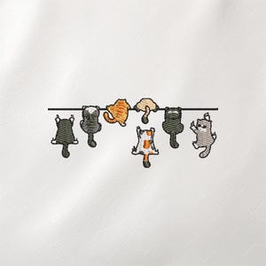 May include: Embroidered design featuring seven stylized cats hanging from a black line. The cats are in various colors, including black, gray, orange, and calico. The design is on a white background, creating a simple yet charming aesthetic.