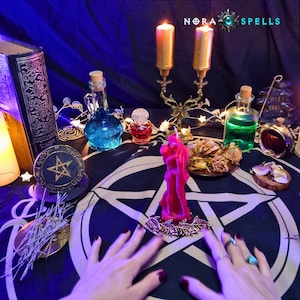 May include: A dark blue altar cloth with a white pentagram features a pink candle shaped like two figures embracing, a gold candelabra with two lit candles, glass bottles with blue and green liquids, a small red bottle, and a book labeled NORA SPELLS.