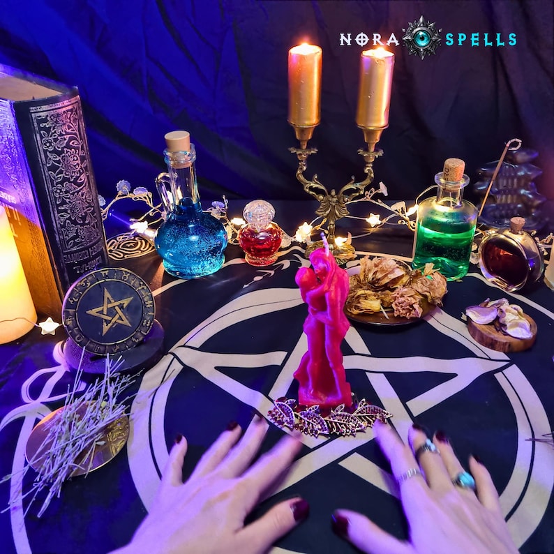 May include: A dark blue altar cloth with a white pentagram features a pink candle shaped like two figures embracing, a gold candelabra with two lit candles, glass bottles with blue and green liquids, a small red bottle, and a book labeled NORA SPELLS.