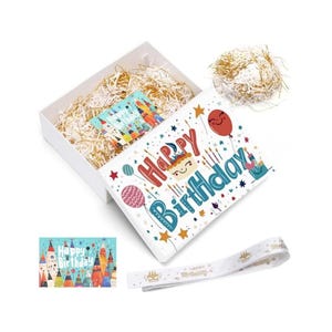 May include: A white gift box filled with gold shredded paper, a greeting card, and a ribbon. The box lid features the words "Happy Birthday" in red and blue, with colorful balloons and stars. A separate card and ribbon are also visible.