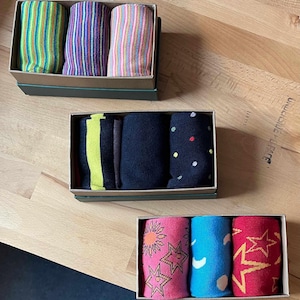 May include: Three sets of socks in boxes. The first set has green, purple, and brown striped socks. The second set has black, yellow, and white polka dot socks. The third set has pink, blue, and red socks with a sun, moon, and star design.
