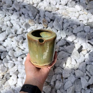 May include: A cylindrical ceramic vessel with a glossy, variegated glaze in shades of sage green and brown. The container features a smooth, rounded rim and a small, circular opening near the top edge, showcasing a reactive finish with earthy tones.
