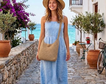 May include: A woman in a light blue dress, straw hat, and woven shoulder bag strolls along a cobbled street. White buildings, potted plants, and the sea form the backdrop. The dress is knee-length, and the bag is a natural beige colour.