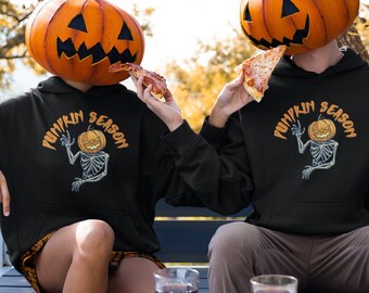 Peut inclure: Deux personnes portant des sweats à capuche noirs avec le texte "PUMPKIN SEASON" et un squelette avec un motif de tête de citrouille. Ils tiennent des parts de pizza et portent des têtes de citrouille. Les sweats à capuche sont noirs.