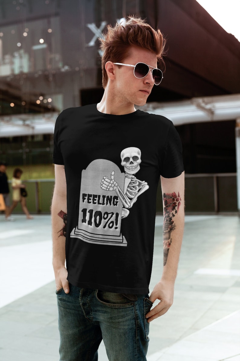 Puede incluir: Camiseta negra con un esqueleto que levanta el pulgar detr&aacute;s de una l&aacute;pida. La l&aacute;pida dice "FEELING 110%!" en texto blanco. La camiseta la lleva una persona con gafas de sol.