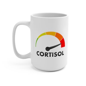 May include: White ceramic mug with a black handle featuring a gauge design. The gauge displays a color gradient from green to red, with a black needle pointing towards the red zone. The word "CORTISOL" is printed in black below the gauge.