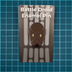 May include: A brown enamel pin featuring a Battle Droid design, with two yellow eye details. The pin is on a card with the text "Battle Droid Enamel Pin" and a gray background with vertical lines. The pin is a collectible item.