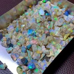 May include: A metal tray filled with a variety of small, iridescent opal gemstones. The opals are various shades of white, blue, green, and pink.
