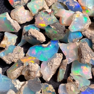 May include: A collection of raw, unpolished opal stones featuring a milky white base with vibrant flashes of iridescent green, blue, orange, and pink color play. Each stone displays a rough, natural texture with earthy, greyish rock matrix.
