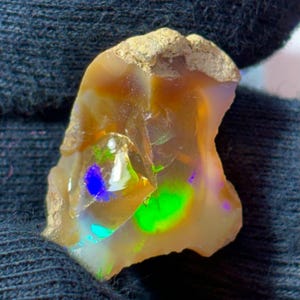 Puede incluir: Una piedra preciosa de ópalo en bruto y sin pulir que presenta un cuerpo translúcido de color miel con destellos vibrantes de verde neón y azul intenso. La piedra tiene una forma orgánica irregular con una matriz rugosa y texturizada en el borde superior.