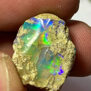 Puede incluir: Una piedra preciosa de ópalo en bruto sin pulir que presenta una matriz exterior rugosa de color arena que rodea un centro iridiscente vibrante que muestra destellos de luz verde, azul y amarilla brillante en su superficie irregular.