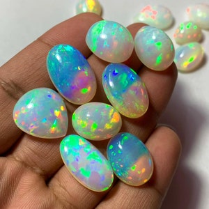 May include: A collection of oval-shaped, iridescent opal cabochons in various shades of white, green, blue, and orange. The opals are displayed on a hand.