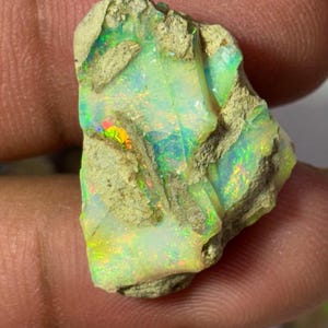 May include: A rough opal gemstone with a matrix of tan rock. The opal displays flashes of green, blue, and red iridescence. The stone has a natural, unpolished appearance, showcasing its raw beauty.
