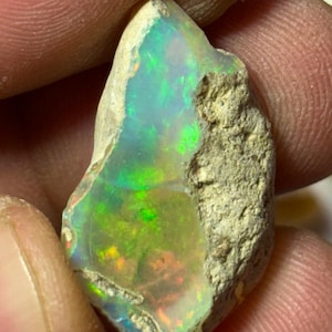 May include: A rough opal gemstone with a vibrant display of color. The stone exhibits a play of light with flashes of green, yellow, and orange. The opal is set against a rough, beige matrix, showcasing its natural, unpolished state.