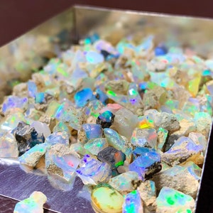 May include: A close-up of a silver tray filled with rough, iridescent opal gemstones. The opals are various sizes and shapes, with a mix of white, blue, green, and pink hues.