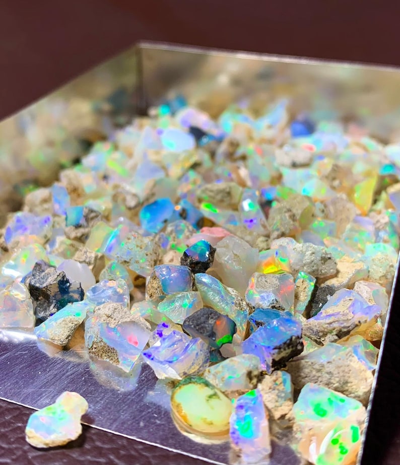 May include: A close-up of a silver tray filled with rough, iridescent opal gemstones. The opals are various sizes and shapes, with a mix of white, blue, green, and pink hues.