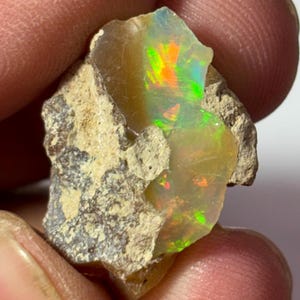 Könnte beinhalten: Ein roher, ungeschliffener Opal-Edelstein mit einem lebendigen Farbspiel. Der Opal zeigt grüne, orange und gelbe Blitze in einer durchscheinenden, milchigen Basis. Der Stein ist in einer Matrix aus beigem Gestein eingebettet.