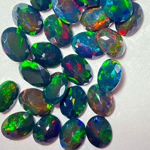 May include: A collection of oval-shaped, faceted black opals featuring a dark base color with vibrant flashes of green, red, and blue light. Each gemstone displays a polished surface with multiple geometric facets that reflect light across the stones.
