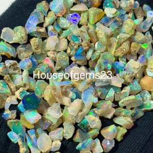 May include: A collection of raw opal gemstones, displaying a variety of colors including blue, green, and yellow. The opals have a rough, natural texture and are scattered across a dark surface. The text "Houseofgems23" is visible.