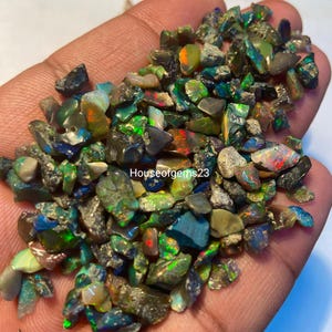 May include: A handful of rough, colorful opal gemstones. The opals display a variety of colors, including green, blue, orange, and red, with a shimmering, iridescent quality. The text "Houseofgems23" is visible.