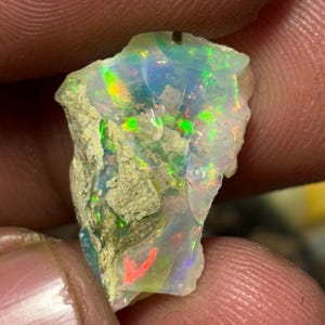 Puede incluir: Una piedra preciosa de ópalo en bruto y sin pulir con una base blanca lechosa translúcida y destellos vibrantes de juego de colores iridiscentes en verde, azul y rojo. La piedra tiene una forma irregular y dentada con una matriz rocosa rugosa.