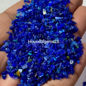 May include: A collection of small, crushed, deep blue opal fragments with iridescent flashes of green, yellow, and white. The text Houseofgems23 is overlaid in white across the center of the pile of vibrant, irregularly shaped gemstone pieces.