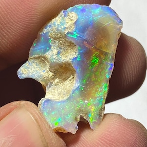 Op de afbeelding: Een ruwe opaal edelsteen met een levendige weergave van kleuren. De opaal heeft flitsen van blauwe, groene en gele tinten, met een getextureerd, natuurlijk oppervlak. De edelsteen wordt tussen vingers gehouden en toont zijn unieke iriserende glans.